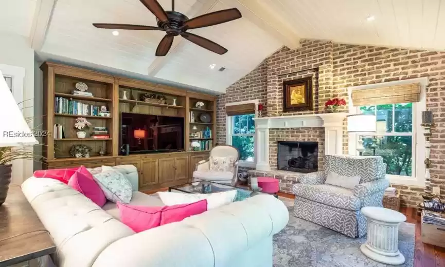 Additional living area featuring a brick fireplace, ceiling fan, heart pine floors, brick wall, and vaulted ceiling with beams.
