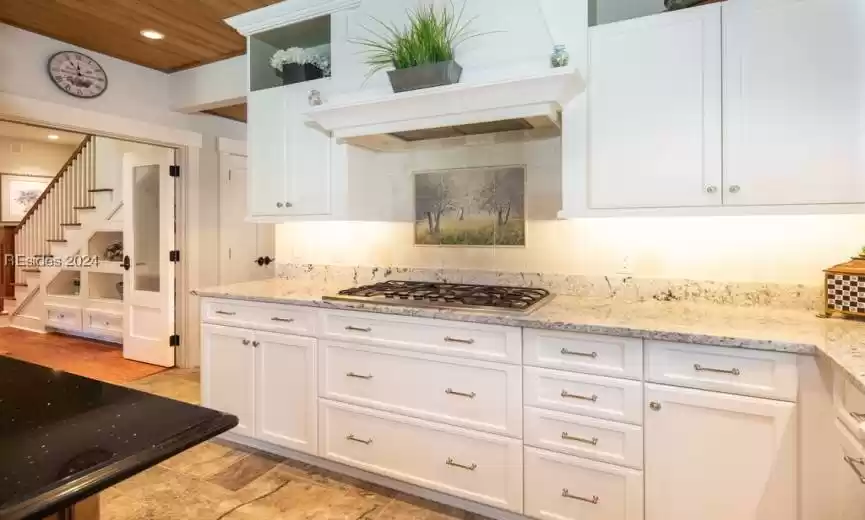 Kitchen with custom exhaust hood, stainless steel gas cooktop, light stone counters, and tasteful backsplash. Cabinets feature glass panels and floating shelves.