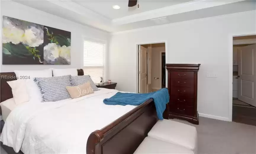 Primary Bedroom view towards the connected bathroom and door to the hallway which has a linen closet and the laundry room.  The bedroom has a ceiling fan, can lights, crown molding, carpet, and a crown molding in the tray ceiling