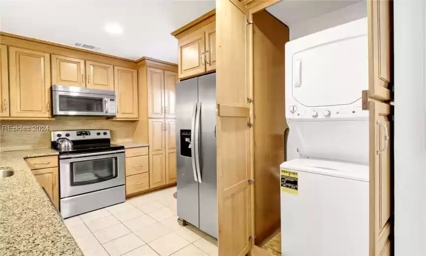 Kitchen with light brown cabinets, light stone counters, stainless steel appliances, stacked washing maching and dryer, and light tile flooring