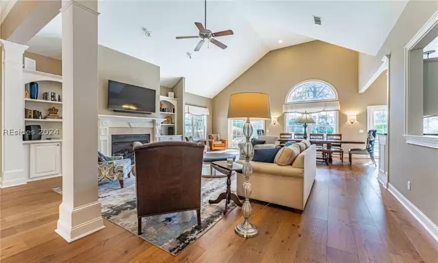 Living room with ceiling fan, a tile fireplace, built in features, ornate columns, and light wood flooring