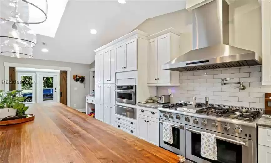 Kitchen featuring wall chimney exhaust hood, wood counters, white cabinetry, backsplash, and double oven range