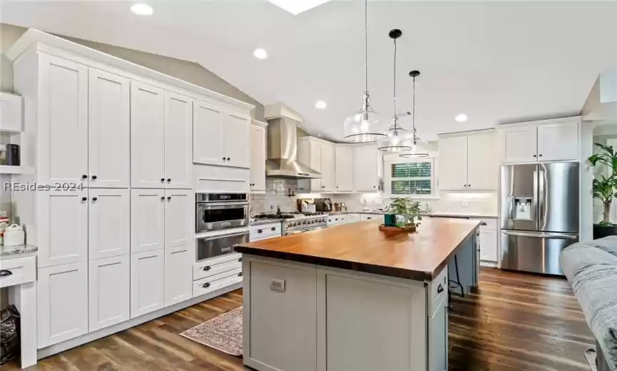 Kitchen featuring a kitchen island, backsplash, stainless steel appliances, wall chimney range hood, and wood counters