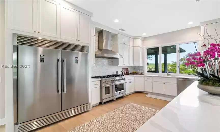 Kitchen featuring high quality appliances, light wood-type flooring, wall chimney exhaust hood, and light stone counters